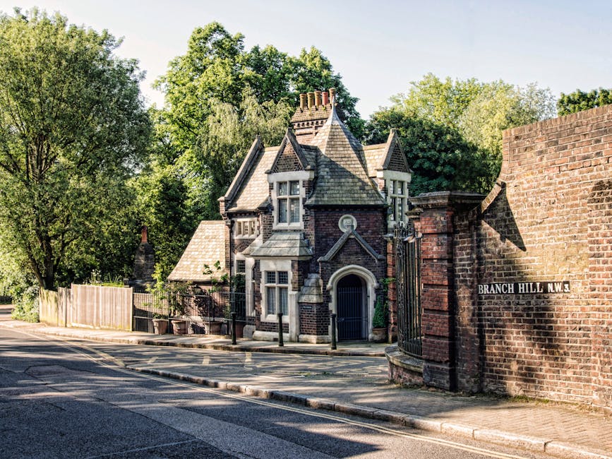 A narrow residential street scene showing a large, historic brick house with a steeply pitched roof, small windows, and decorative stonework, partially obscured by green-leaved trees and a wooden fence. To the right, a brick wall with a street sign reading 'Branch Hill N.W.3' runs alongside the sidewalk, which is paved with concrete slabs. The road in the foreground is paved with asphalt and features a yellow dashed centre line. The scene is illuminated by bright, natural daylight, suggesting a clear, sunny day. This image relates to house removals or home relocation services provided by Man and Van Chiswick, highlighting the exterior environment involved in furniture transport and packing and moving logistics.