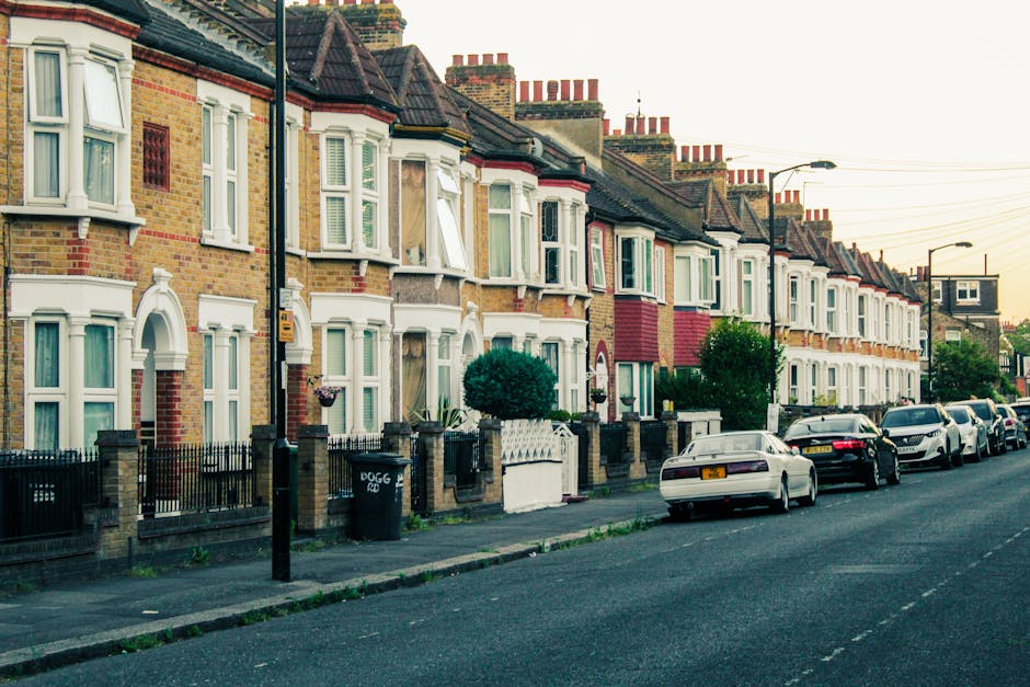 A quiet residential street in Chiswick lined with traditional brick terraced houses featuring bay windows and chimneys. The pavement is bordered by well-maintained greenery, including bushes, flowering plants, and large leafy trees casting shade over the road. A tall street lamp stands beside the pavement, and the sky overhead is clear with a bright blue hue. In the context of a house removal or relocation, the scene depicts a typical suburban setting where furniture and packing materials might be transported to or from a home. Man and Van Chiswick's services could involve loading or unloading household items in this area, with moving equipment such as trolleys or blankets potentially visible during the process, although they are not present in this particular image.