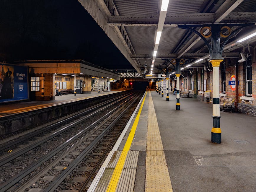 A view of Turnham Green station platform at night, featuring multiple parallel train tracks with dark ambient lighting overhead. The platform has a yellow tactile paving strip along its edge for safety, and is lined with evenly spaced decorative black and yellow bollards surrounding support columns. On the left side, there is a small waiting area with a bench and a person sitting. In the distance, the platform continues with additional seating, lighting, and a covered waiting area. The station walls are brick with posters and signs, and a nameboard with the London Underground roundel symbol is visible on the right side. The scene provides a clear view of the station's structural details, lighting, and signage, conveying a typical British railway station environment suitable for home relocation or moving logistics context, as handled by [COMPANY_NAME] and their removals services.