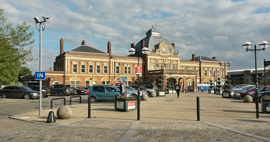 A wide-view image of Turnham Green station in Chiswick, showing an ornate historic brick building with decorative architectural details, a central clock, and a prominent dome at the top. In front of the station, there is a paved area with black metal bollards, street lamps, and planters filled with flowers. The foreground features a line of parked cars, including small and medium-sized vehicles, adjacent to the station entrance. The scene is illuminated by daylight, with a partly cloudy sky overhead, and a few pedestrians walking near the entrance. This setting illustrates the typical environment for home relocation or furniture transport services provided by Man and Van Chiswick, with vehicles potentially loading or unloading near the station as part of a packing and moving process.