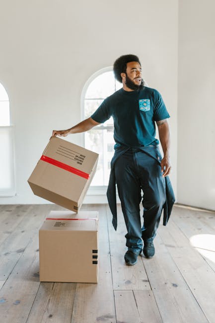 A man with dark skin and an afro hairstyle is inside a bright, unfurnished room with a large arched window that allows natural light to fill the space. He is dressed in a blue t-shirt and dark trousers, holding a cardboard box wrapped with clear packing tape and marked with a red stripe, indicating it is packed for moving. In front of him on the wooden floor are two additional boxes, one aligned beneath the other, with the man appearing to be in the process of loading or stacking them. The room's minimal furnishings emphasize the focus on packing and loading activities typical of home relocation services. The setting, natural daylight, and the man's stance suggest a professional move, with the environment prepared for furniture transport, packing and moving tasks, or house removals. The overall scene subtly highlights the logistics involved in house removals, as would be managed by Man and Van Chiswick, featured on the page about W4 postcode removals costs.
